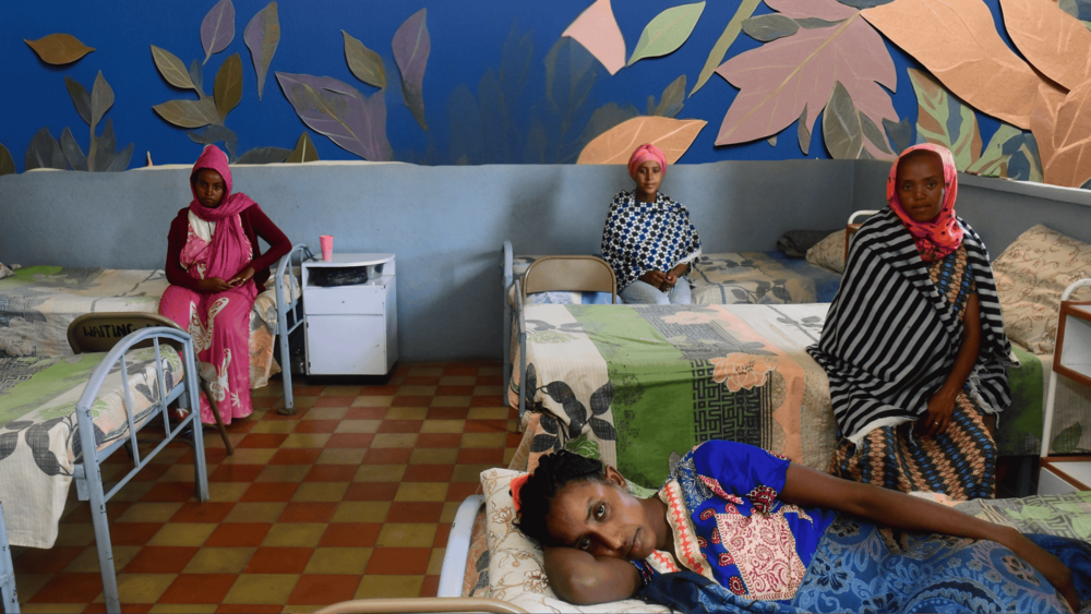 Three women on beds spread out on a hospital bedroom in Eritrea. They are looking at the camera, with tranquil eyes.